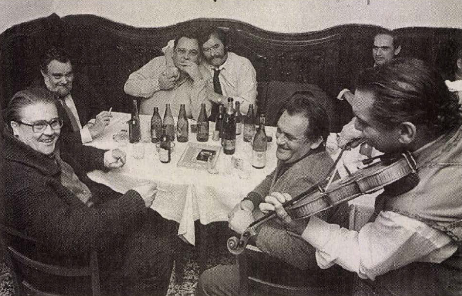 Csukás István barátaival ül egy asztal körül, egyik férfi hegedül