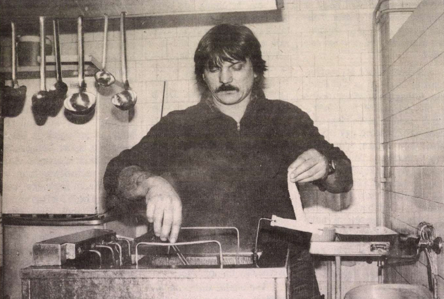 egy bajuszos férfi áll a konyhában, kezében eszköz
